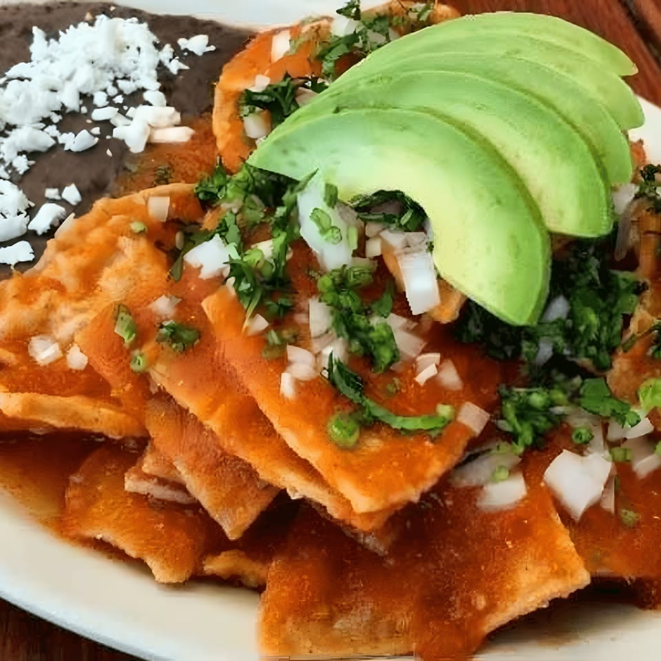 Chilaquiles with red sauce, topped with avocado slices, diced onions, cilantro, and a side of refried beans.